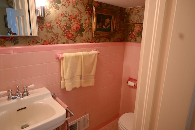 half bathroom featuring a sink, visible vents, wainscoting, and wallpapered walls