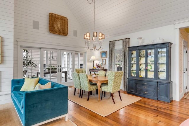 dining space with high vaulted ceiling, wooden walls, a chandelier, light wood-type flooring, and french doors