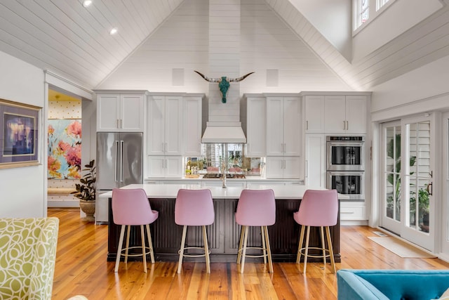 kitchen with appliances with stainless steel finishes, high vaulted ceiling, an island with sink, a kitchen breakfast bar, and custom range hood