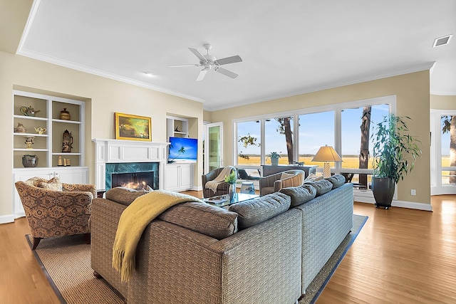 living room featuring built in features, ornamental molding, ceiling fan, a premium fireplace, and light hardwood / wood-style flooring