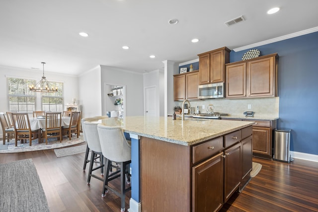 kitchen with visible vents, stainless steel microwave, a sink, a kitchen island with sink, and backsplash