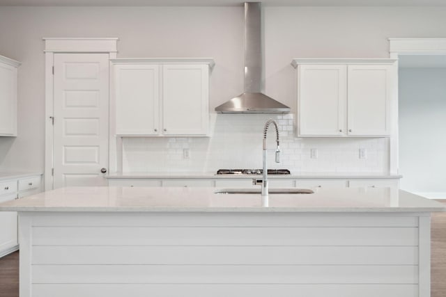 kitchen featuring white cabinetry, wall chimney range hood, a kitchen island with sink, and light stone countertops