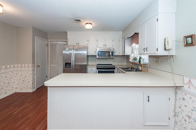 kitchen featuring dark wood-style flooring, stainless steel appliances, a sink, a peninsula, and wallpapered walls