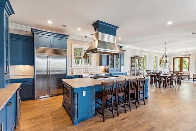 kitchen featuring blue cabinets, island exhaust hood, built in appliances, ornamental molding, and a breakfast bar