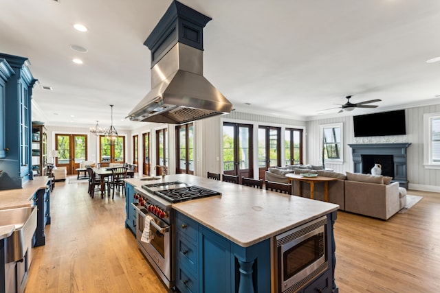 kitchen with ceiling fan with notable chandelier, a fireplace, island range hood, appliances with stainless steel finishes, and blue cabinetry