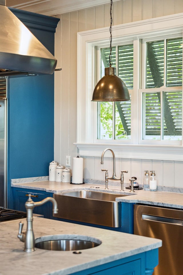 interior space with wall chimney range hood, blue cabinetry, stainless steel dishwasher, a sink, and light stone counters