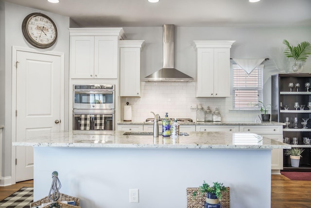 kitchen featuring double oven, wall chimney range hood, a sink, white cabinetry, and decorative backsplash