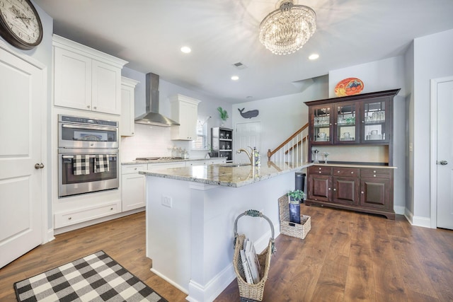 kitchen with wall chimney exhaust hood, appliances with stainless steel finishes, dark wood-type flooring, a chandelier, and white cabinetry