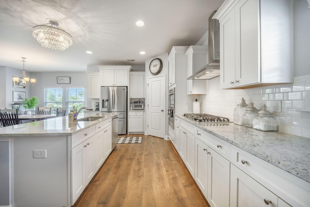 kitchen with a chandelier, wall chimney exhaust hood, appliances with stainless steel finishes, a sink, and white cabinets