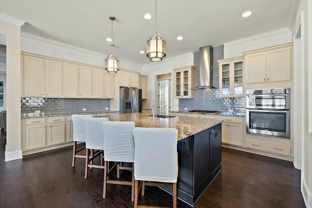 kitchen featuring cream cabinetry, a center island with sink, a sink, stainless steel appliances, and wall chimney range hood