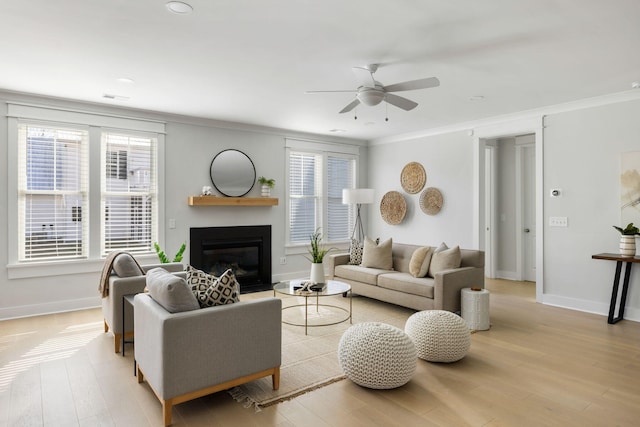 living area with crown molding, light wood finished floors, a glass covered fireplace, ceiling fan, and baseboards