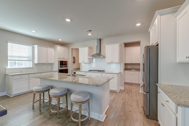 kitchen with an island with sink, wall chimney range hood, appliances with stainless steel finishes, and white cabinets