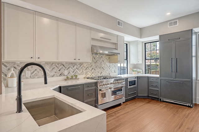 kitchen featuring wall chimney range hood, light stone counters, gray cabinets, high end appliances, and a sink