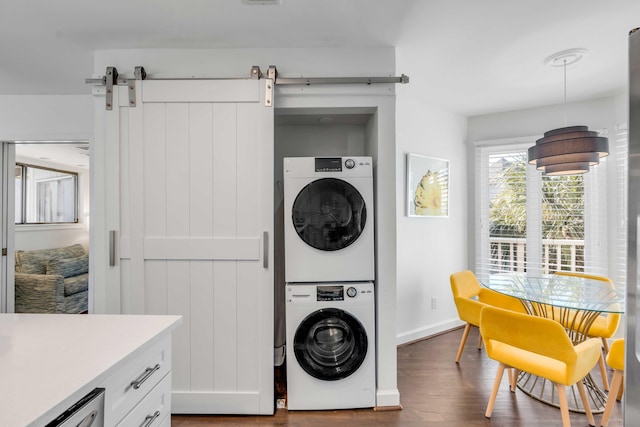 clothes washing area with baseboards, a barn door, laundry area, stacked washing maching and dryer, and dark wood-style floors