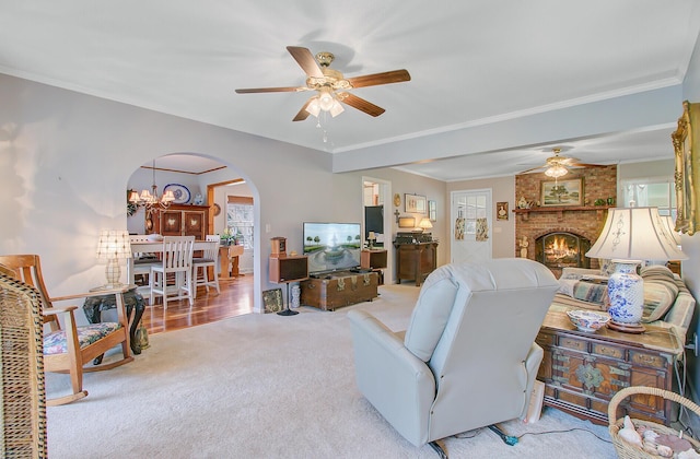 carpeted living room featuring arched walkways, crown molding, ceiling fan with notable chandelier, and a brick fireplace