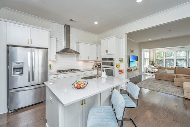 kitchen with wall chimney exhaust hood, stainless steel appliances, a kitchen island with sink, white cabinetry, and sink