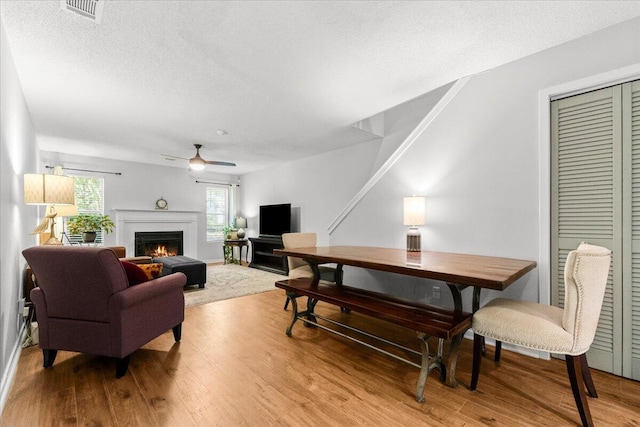 living room with wood finished floors, a textured ceiling, a warm lit fireplace, ceiling fan, and visible vents