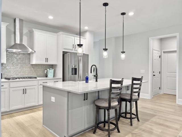 kitchen with appliances with stainless steel finishes, an island with sink, white cabinets, hanging light fixtures, and wall chimney range hood