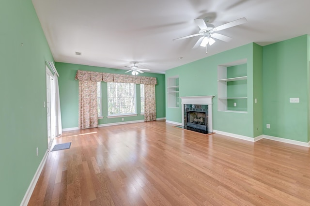 unfurnished living room featuring built in shelves, light hardwood / wood-style floors, a premium fireplace, and ceiling fan