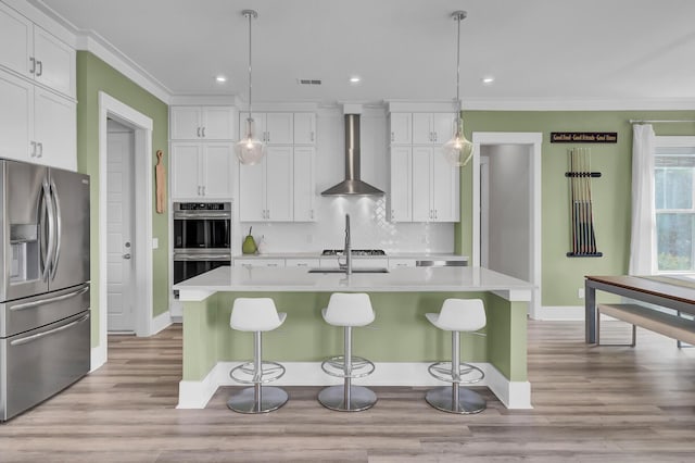 kitchen with an island with sink, appliances with stainless steel finishes, light countertops, wall chimney range hood, and white cabinetry