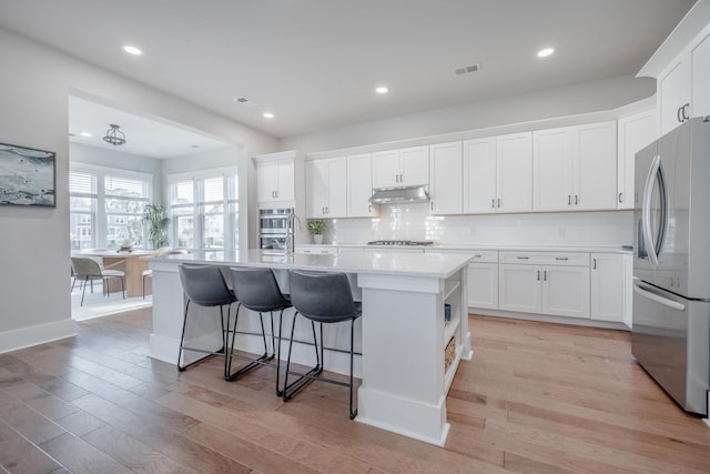 kitchen with white cabinets, a kitchen island with sink, light countertops, under cabinet range hood, and appliances with stainless steel finishes