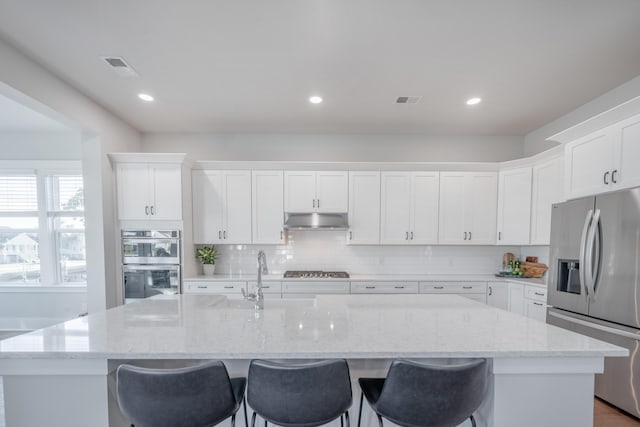 kitchen with a sink, an island with sink, appliances with stainless steel finishes, white cabinetry, and under cabinet range hood