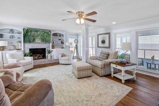 living area featuring built in shelves, a fireplace, wood finished floors, decorative columns, and crown molding