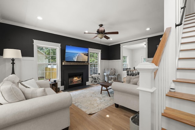 living area featuring stairway, ceiling fan, a fireplace with flush hearth, ornamental molding, and a wainscoted wall