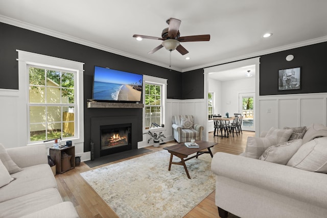 living room featuring ceiling fan, plenty of natural light, a fireplace with flush hearth, wainscoting, and crown molding