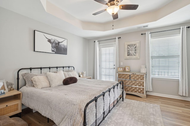 bedroom with ceiling fan, a tray ceiling, light hardwood / wood-style floors, and multiple windows