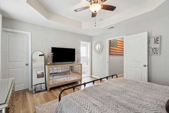 bedroom featuring ceiling fan, connected bathroom, light hardwood / wood-style floors, and a tray ceiling