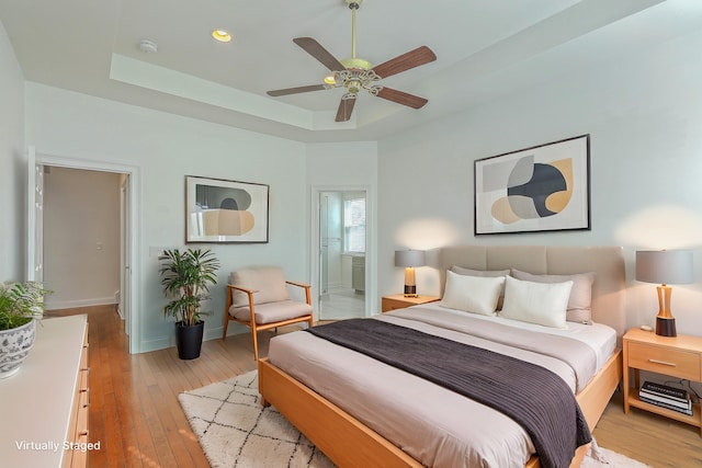 bedroom featuring light wood-type flooring, ceiling fan, connected bathroom, and a tray ceiling