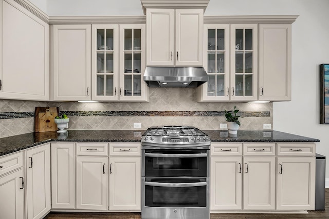kitchen featuring double oven range, under cabinet range hood, decorative backsplash, dark stone counters, and glass insert cabinets