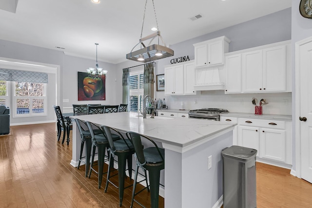 kitchen featuring white cabinets, decorative light fixtures, premium range hood, a center island with sink, and stainless steel range with gas stovetop