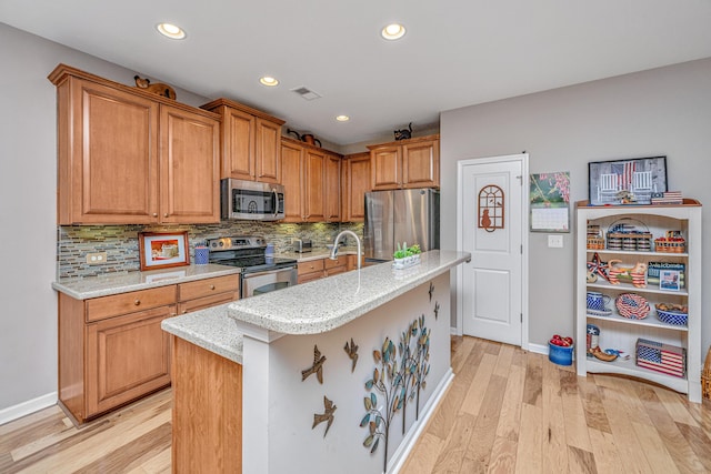 kitchen with appliances with stainless steel finishes, light wood-type flooring, tasteful backsplash, light stone counters, and a kitchen island with sink