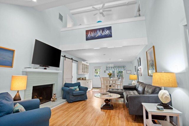 living room with ceiling fan, a barn door, high vaulted ceiling, light wood-type flooring, and a brick fireplace