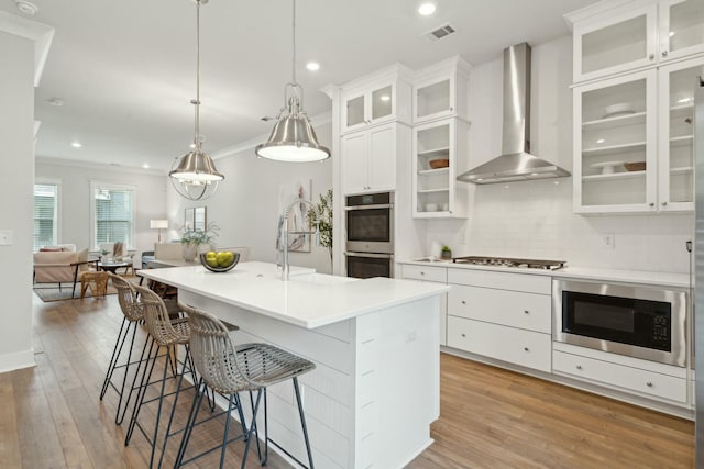 kitchen featuring appliances with stainless steel finishes, glass insert cabinets, light countertops, wall chimney range hood, and white cabinetry