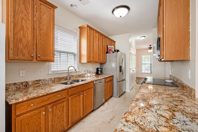 kitchen with a sink, light stone counters, ceiling fan, brown cabinetry, and appliances with stainless steel finishes