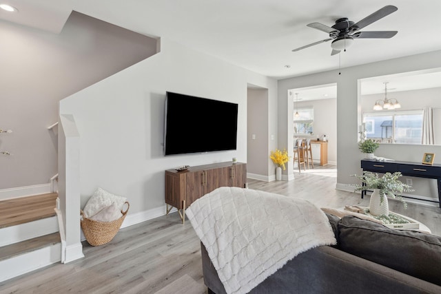 living room with ceiling fan with notable chandelier, light wood-type flooring, baseboards, and stairs