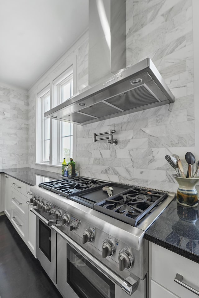kitchen featuring decorative backsplash, wall chimney exhaust hood, white cabinets, range with two ovens, and dark stone countertops