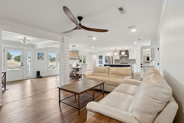 living room with ceiling fan with notable chandelier, decorative columns, sink, crown molding, and light hardwood / wood-style flooring