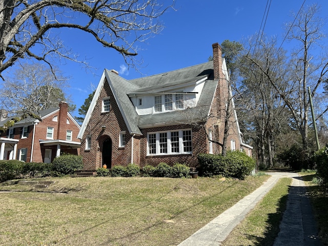 english style home featuring brick siding, a chimney, a front yard, and a shingled roof