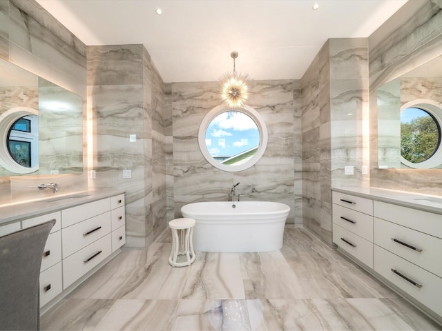 full bath featuring marble finish floor, tile walls, two vanities, a soaking tub, and a sink