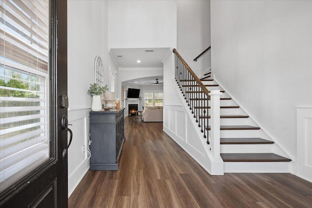 entryway featuring ornamental molding, a warm lit fireplace, dark wood-style floors, stairway, and ceiling fan