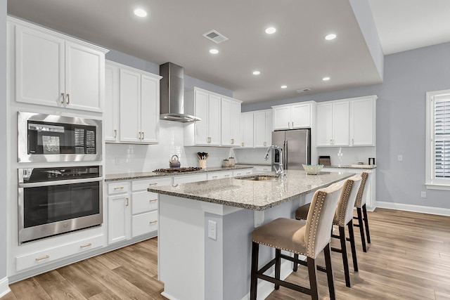 kitchen with sink, a center island with sink, wall chimney range hood, stainless steel appliances, and white cabinets