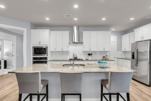 kitchen with wall chimney range hood, appliances with stainless steel finishes, an island with sink, white cabinets, and a kitchen bar