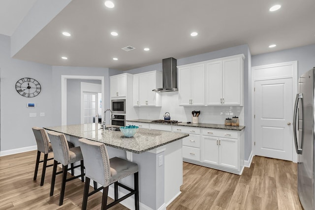 kitchen featuring stone counters, an island with sink, white cabinetry, stainless steel appliances, and wall chimney exhaust hood