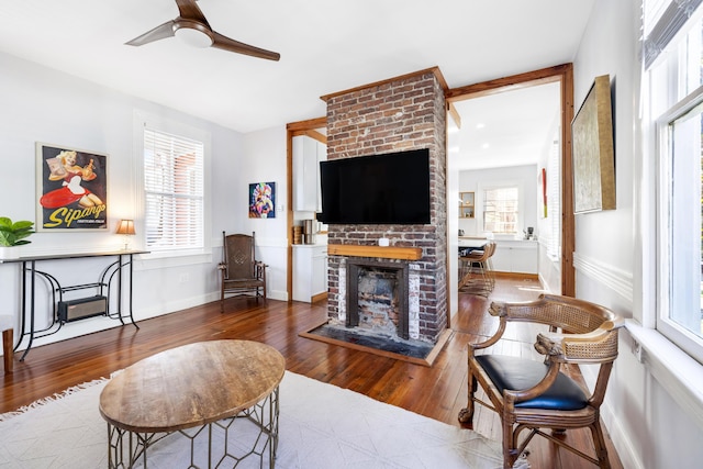 living room with a brick fireplace, ceiling fan, baseboards, and wood finished floors