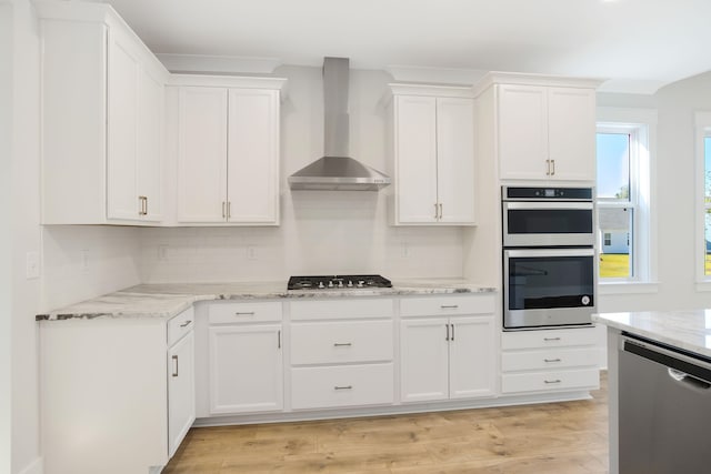 kitchen with wall chimney range hood, appliances with stainless steel finishes, white cabinetry, light stone countertops, and light wood-type flooring