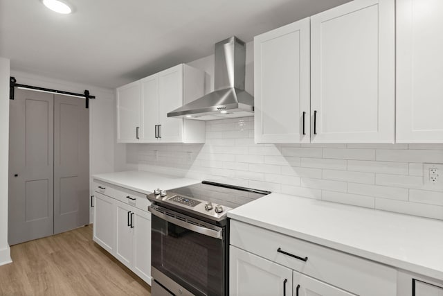 kitchen featuring stainless steel electric range, a barn door, wall chimney range hood, and white cabinetry